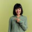 joyful young brunette caucasian girl points at side looking at camera isolated on olive green background with copy space